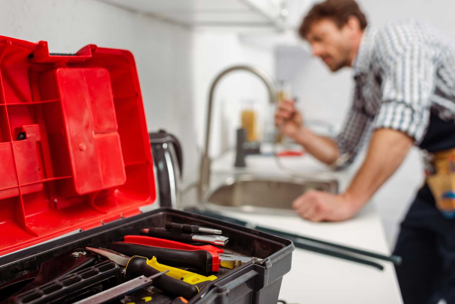Plumber working at a kitchen sink with an open red tool box filled with tools in the foreground